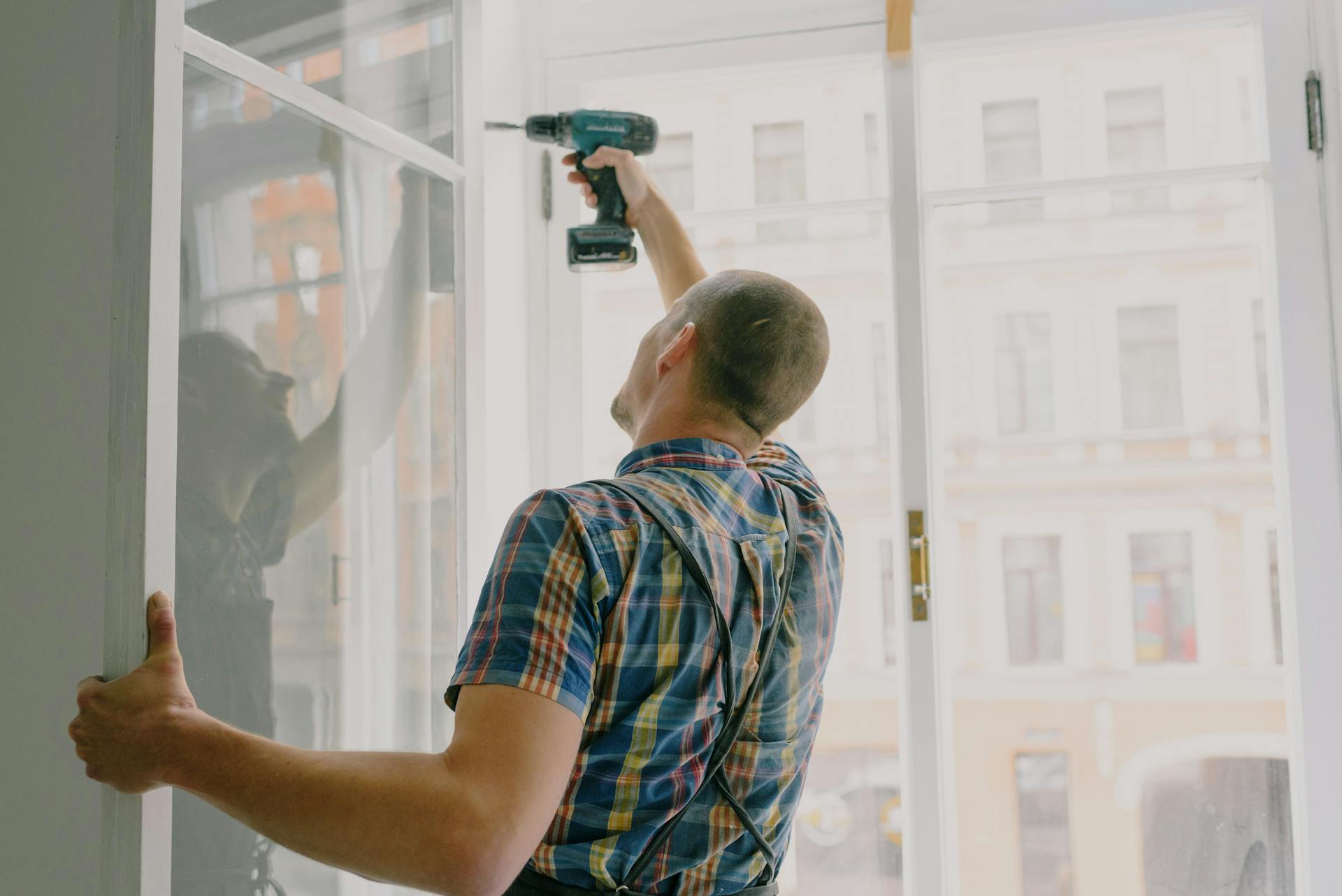 Back view of worker in checkered shirt drilling window frame during renovation process in apartment