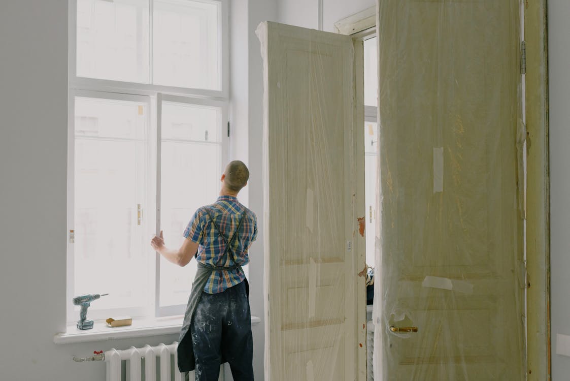 Free Back view of unrecognizable workman in apron mounting window near drilling machine and doors in flat Stock Photo