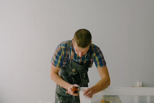 Concentrated anonymous joiner in apron working with wooden plank for furniture in light room