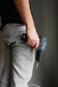 A man holds a cordless drill, ready for work, against a neutral indoor background.