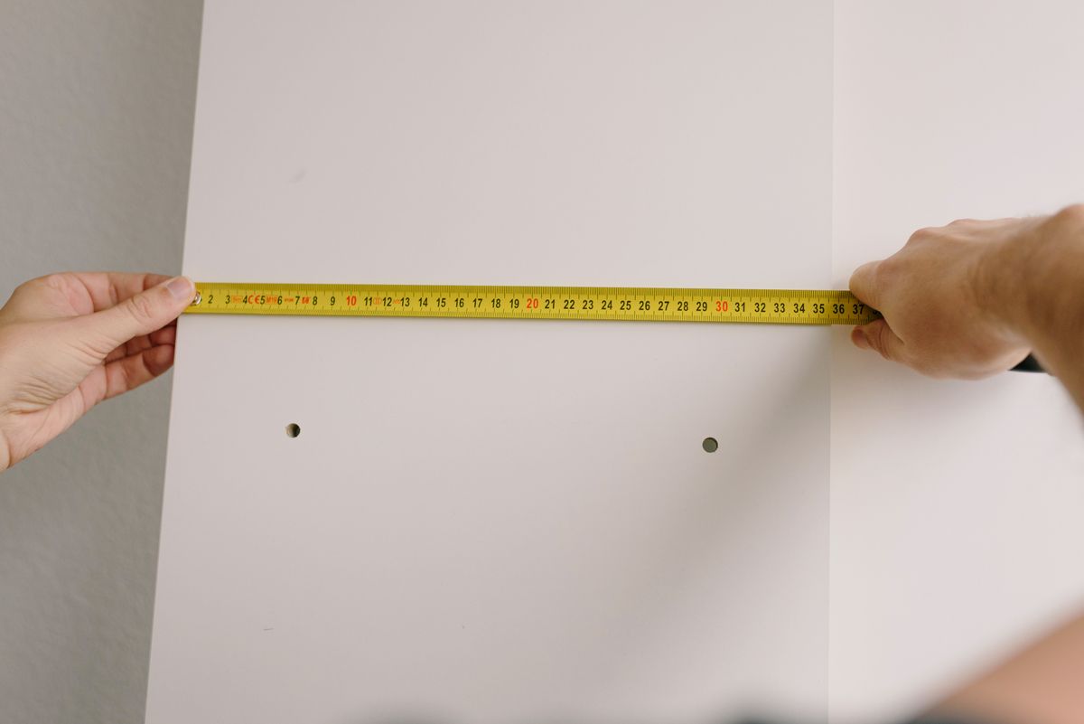 Two people measure a white cabinetry panel with a tape measure; drilled mounting holes are visible in the panel.