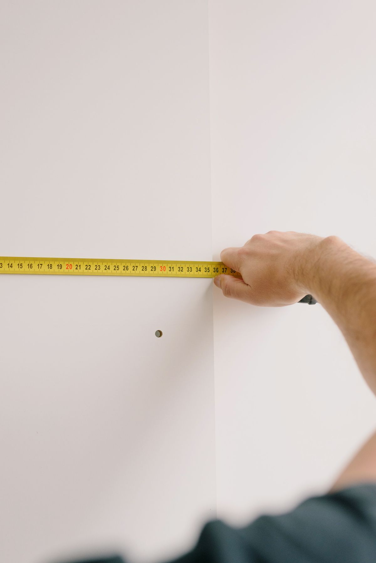 A hand holds a yellow tape measure across a white wall or cabinetry panel, with a small drilled hole visible below.