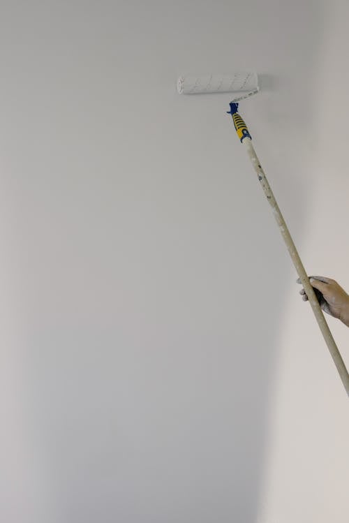 Crop anonymous person holding roller with white paint and painting wall in light apartment