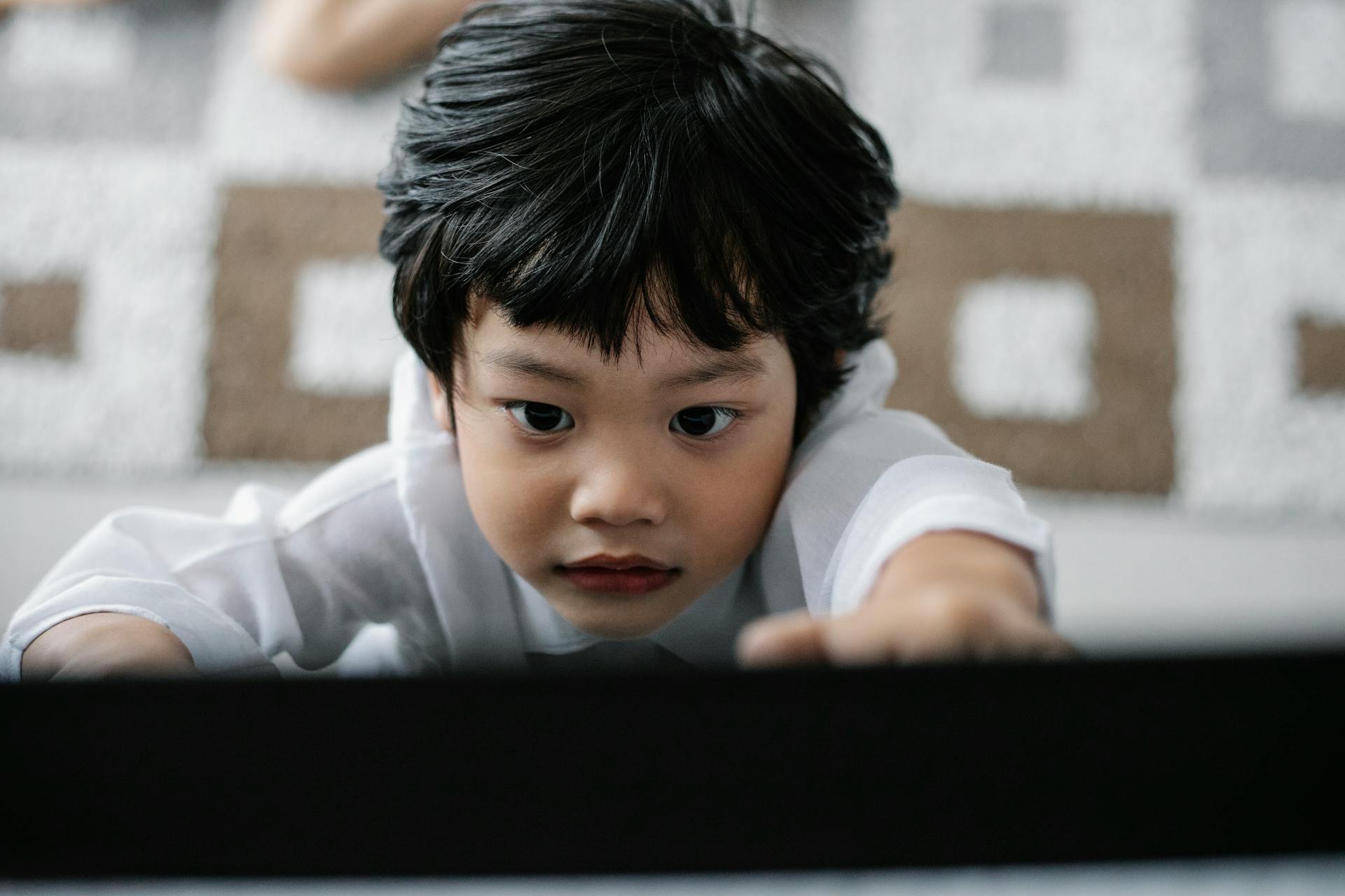 An attentive child with dark hair reaches forward indoors, conveying curiosity and focus.