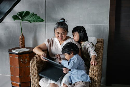 Calm Asian woman sitting with kids and using tablet while spending time together at home