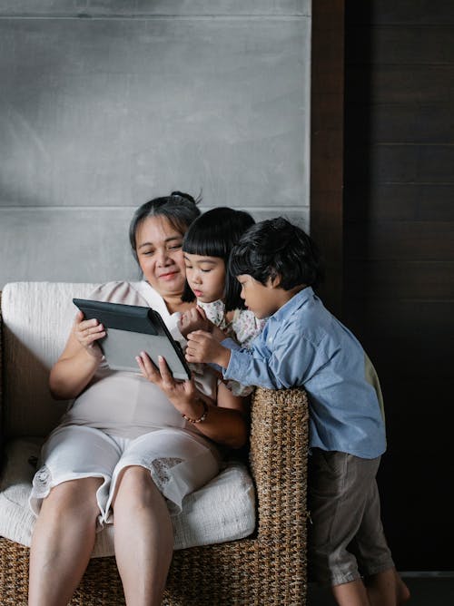 Free Positive Asian woman sitting with children and watching at tablet in cozy light room Stock Photo