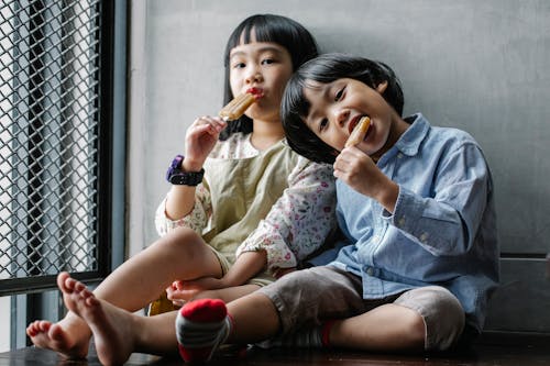 Positive Asian children in casual clothes sitting on floor and eating yummy ice creams