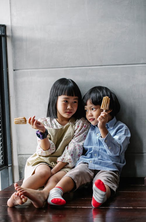 Calm Asian children raising hands with ice creams and sitting on floor at home