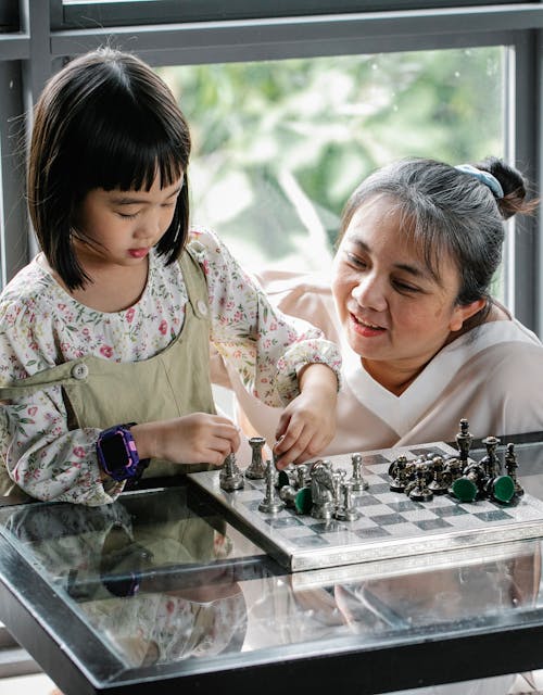 Free Concentrated Asian child standing near glass table and playing chess with mother at home Stock Photo