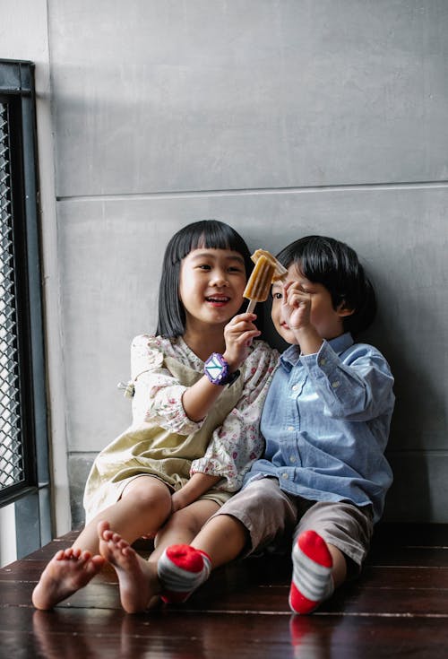 Cute Asian kids clinking ice creams and sitting on floor