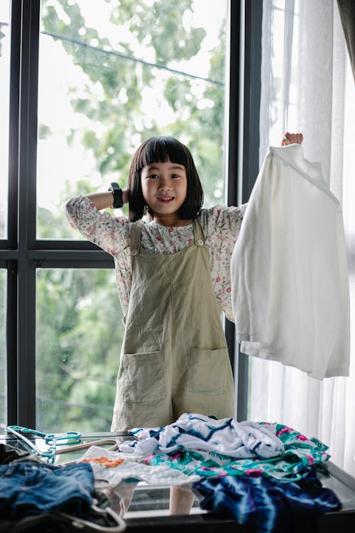Fröhliches Asiatisches Mädchen, Das Kleidung Auf Tisch Gehäuft Sortiert