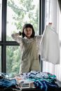 Happy Asian girl in casual wear raising hand with shirt on hanger while sorting clothes heaped on table and looking at camera with smile