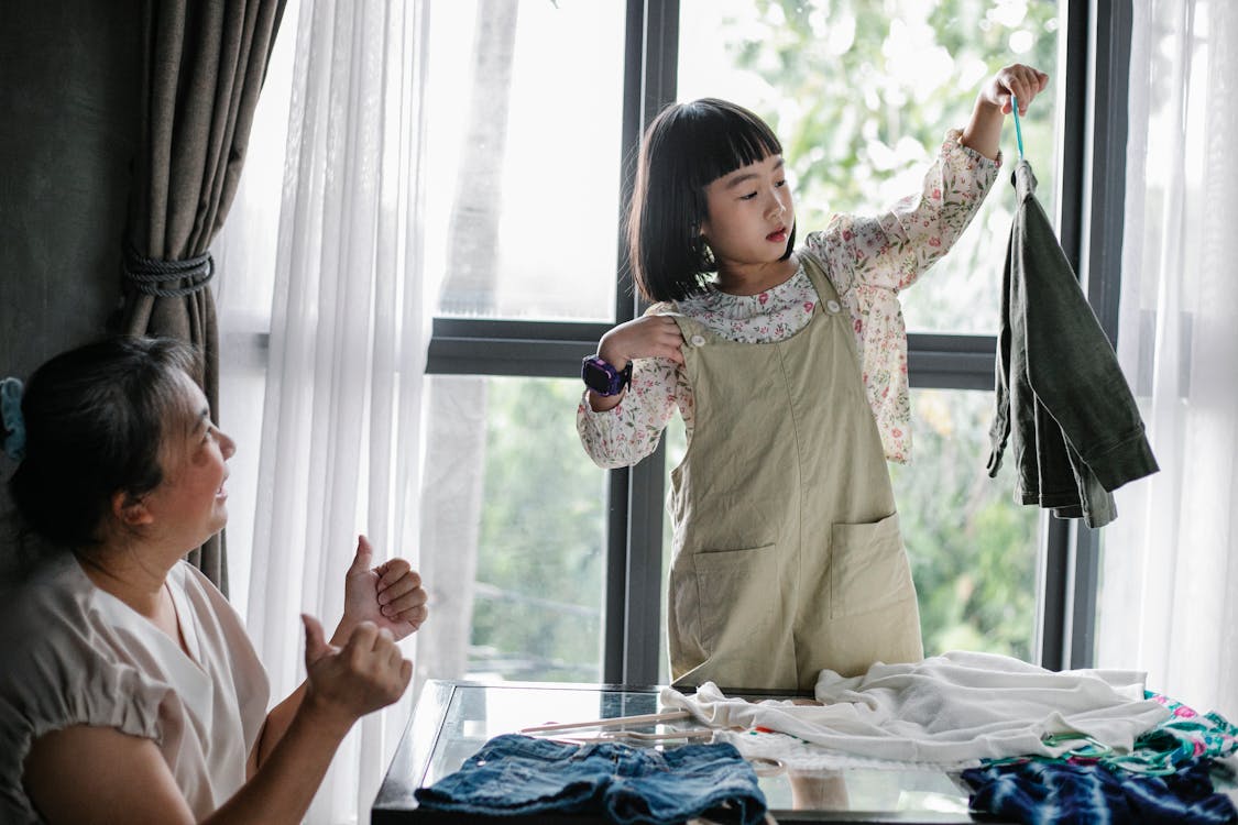 Happy Asian woman showing like gesture while granddaughter organizing clothes on hanger at home