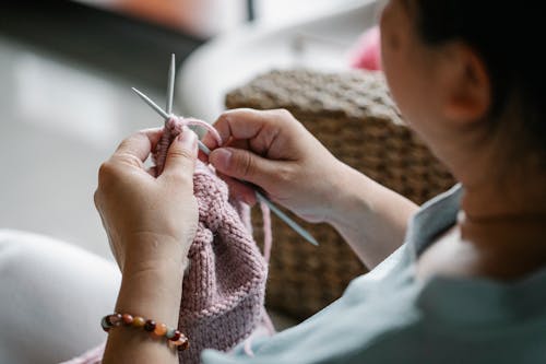 Crop faceless female holding needles and knitting pink cloth while sitting at home