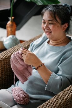 一位中年亚洲妇女坐在家中的柳条扶手椅上，微笑着织毛衣