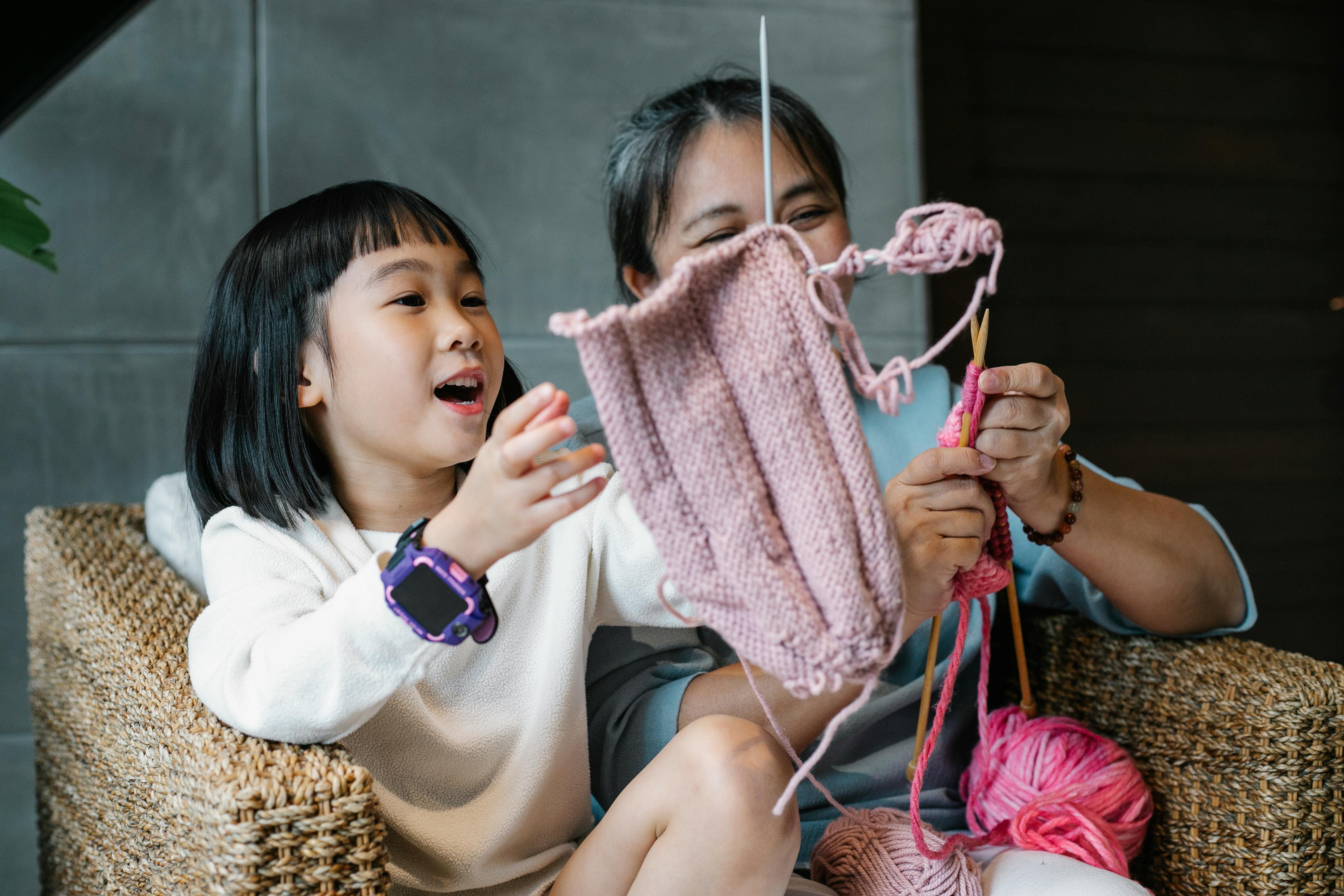 Charming Asian girl with laughing woman in armchair at home knitting together and having fun