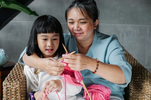 Content mature Asian woman embracing little granddaughter and explaining how to knit with needles