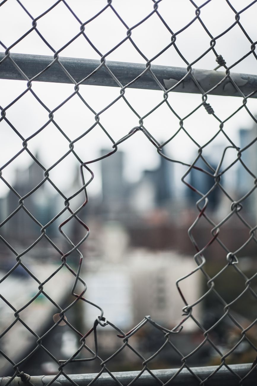 A city skyline viewed through a broken chain link fence, highlighting urban decay.