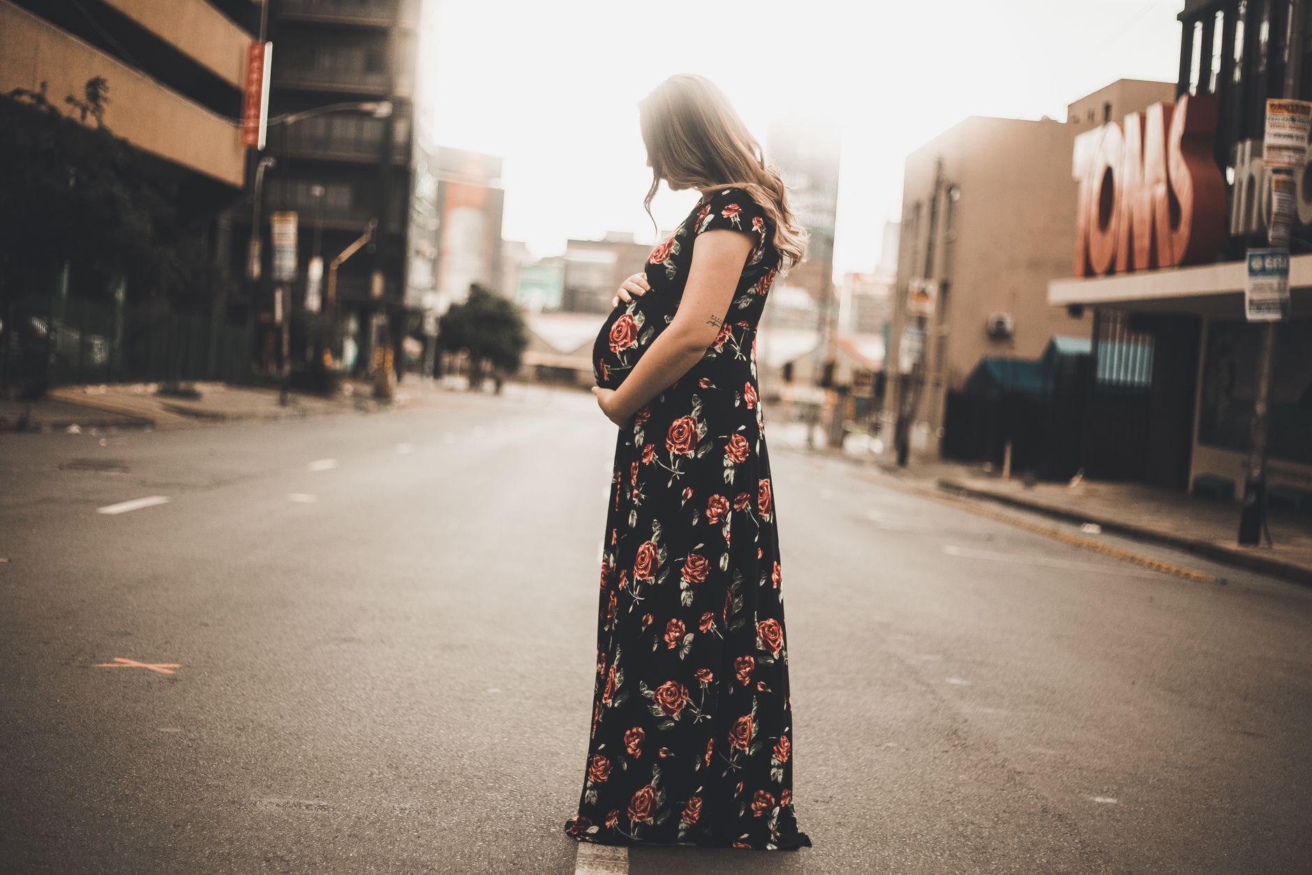 Pregnant woman in floral dress standing on empty city street at sunset.