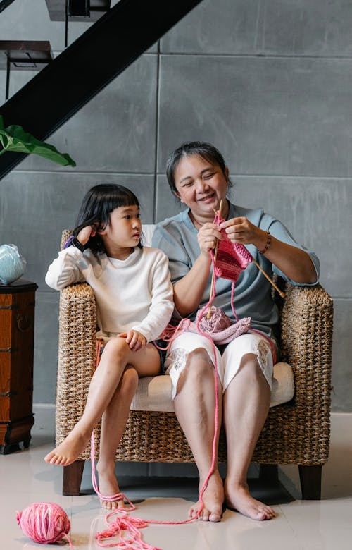 Positive senior Asian female knitting while sitting on armchair with cute granddaughter