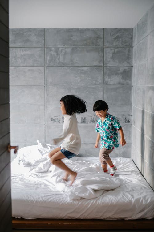 Free Side view of expressive Asian little children jumping on comfy soft bed while spending time together at home Stock Photo