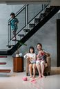 Full length of happy elderly Asian woman sitting on armchair and knitting while spending weekend with cute grandchildren in stylish villa with staircase and modern interior