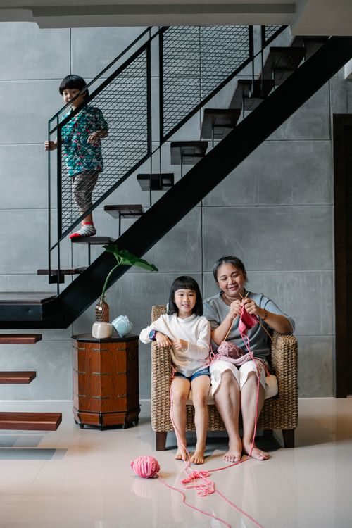 Free Full length of happy elderly Asian woman sitting on armchair and knitting while spending weekend with cute grandchildren in stylish villa with staircase and modern interior Stock Photo