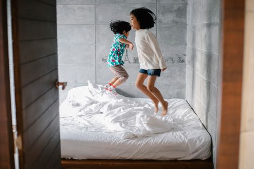 Free Side view of cheerful little Asian sister and brother in casual clothes jumping on bed while having fun together at home Stock Photo