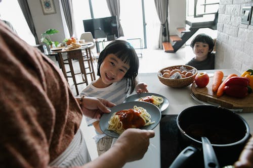 Free High angle of hungry cute little Asian children smiling while waiting for lunch prepared by unrecognizable grandmother standing in kitchen Stock Photo
