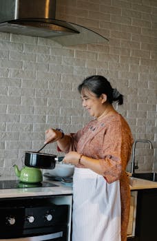 Side view of content elderly ethnic woman in dress and apron stirring hot dish in saucepan and smiling while preparing lunch in stylish kitchen
