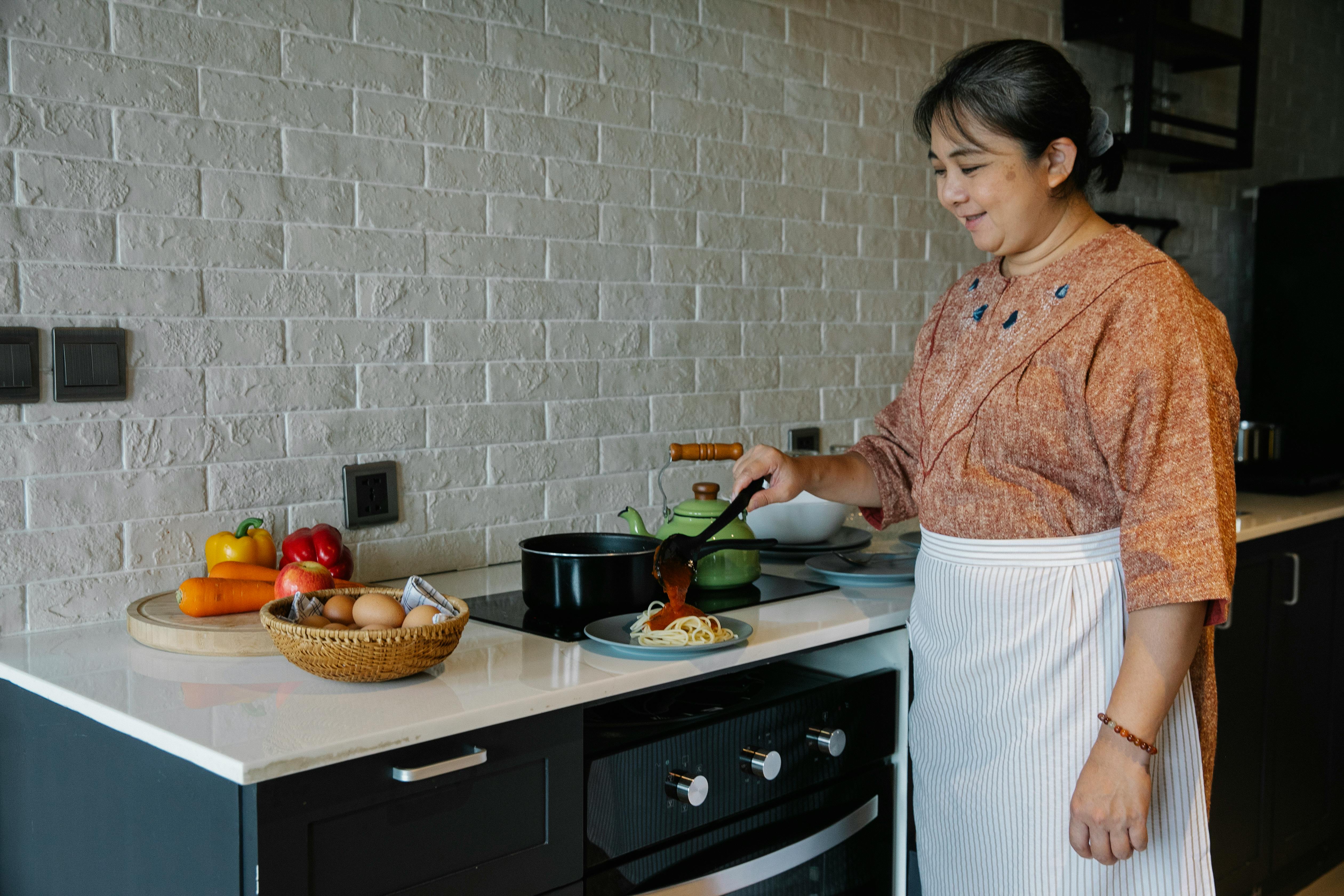 cheerful senior ethnic housewife pouring sauce on pasta while serving dish in kitchen