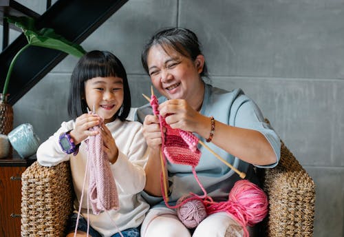 Cheerful little Asian girl with grandmother knitting with needles while sitting together on wicker armchair at home