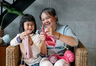Cheerful little Asian girl with grandmother knitting with needles while sitting together on wicker armchair at home