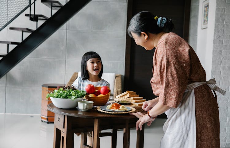 Happy Ethnic Elderly Woman Serving Healthy Lunch For Cute Granddaughter