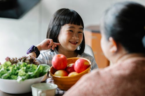 Delighted cute ethnic little girl smiling while sitting at table and eating delicious fresh vegetables and fruits with unrecognizable mother