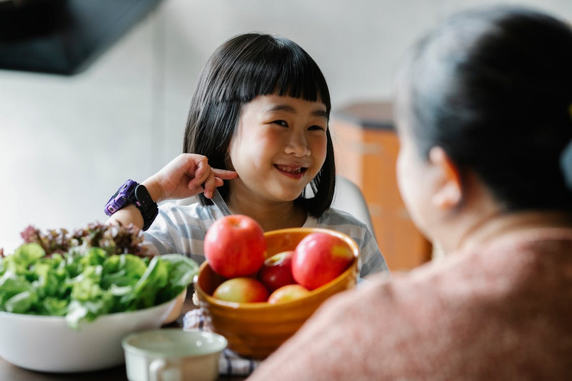Free Delighted cute ethnic little girl smiling while sitting at table and eating delicious fresh vegetables and fruits with unrecognizable mother Stock Photo