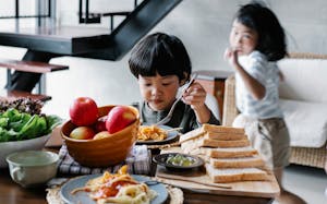 Active little Asian girl running behind brother having lunch in kitchen