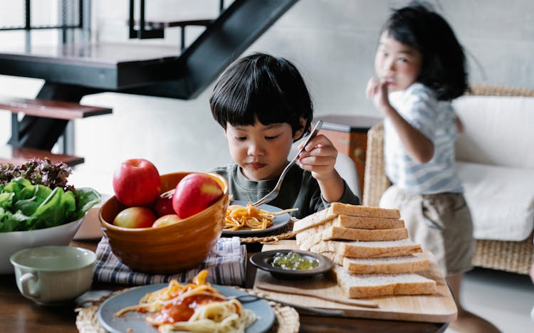 Active Little Asian Girl Running Behind Brother Having Lunch In Kitchen