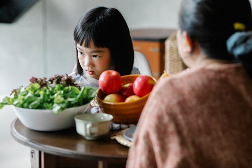 Serious little ethnic girl sitting at table with mother during lunch and looking away