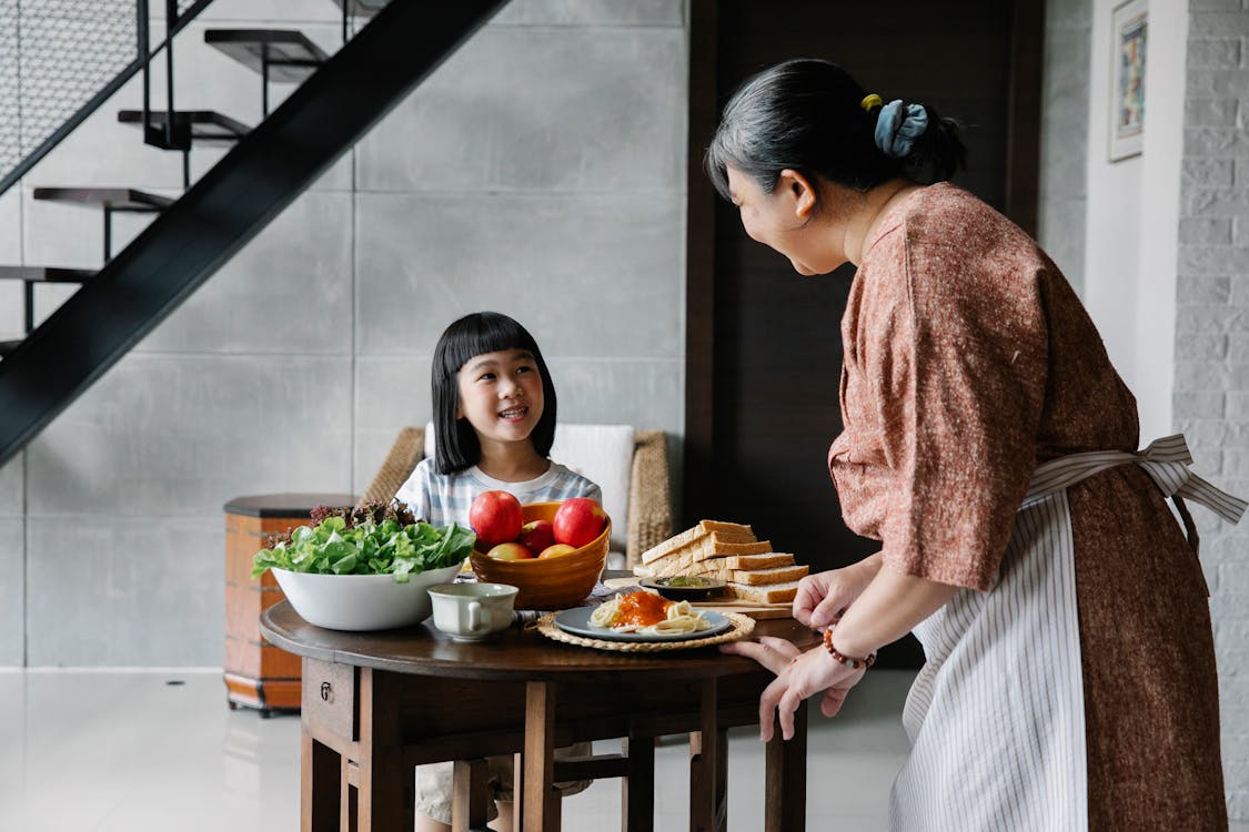 Довольная маленькая азиатская девочка улыбается и разговаривает с бабушкой  во время обеда дома · Бесплатные стоковые фото