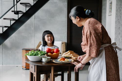Delighted little Asian girl smiling and speaking with grandmother while having lunch at home