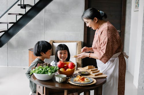 Side view of positive senior Asian female in apron spreading butter on bread while preparing breakfast for funny little grandchildren sitting at round table in cozy kitchen