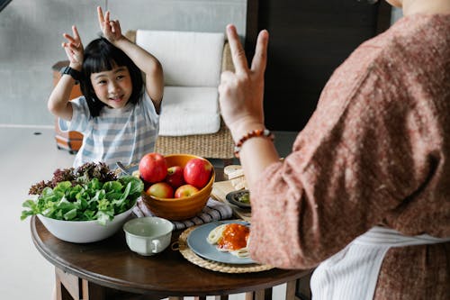 Happy little ethnic girl having fun with faceless mother showing V sign during lunch