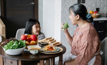 一位戴着围裙和裙子的亚裔老奶奶，正张着嘴吃生菜，与细心可爱的小孙女一起坐在桌旁享用午餐