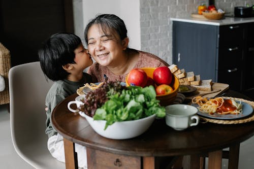 Adorable little ethnic child kissing cheek of smiling grandmother while having lunch together at home