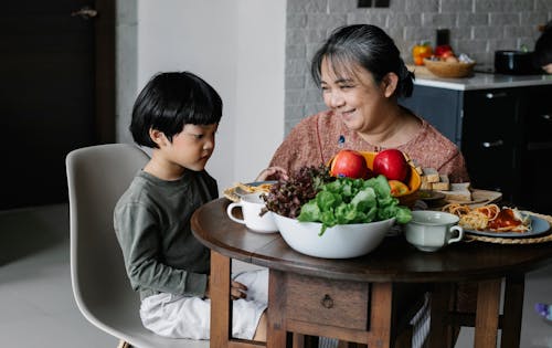 Attentive little Asian boy sitting at table with various delicious dishes prepared by happy grandmother during weekend at home