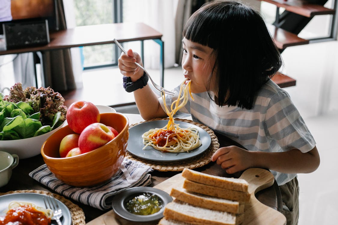 Cute Asian little girl enjoying delicious spaghetti during lunch at home