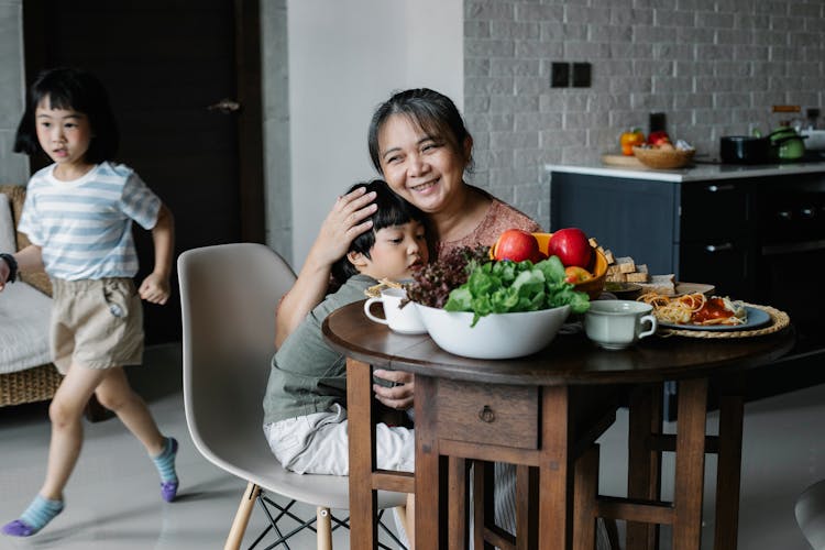 Active Little Asian Girl Running Behind Grandmother And Brother Embracing At Table During Breakfast