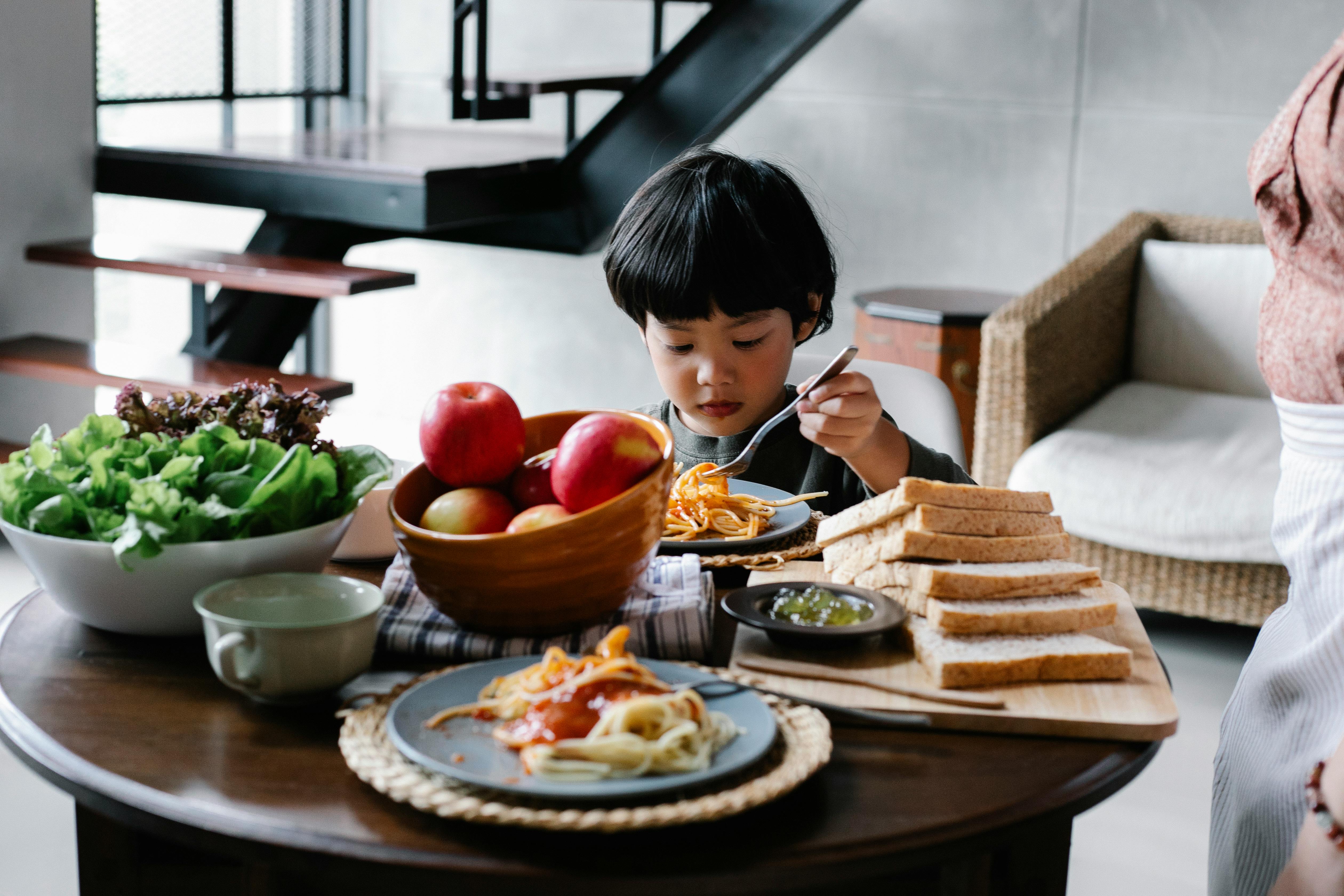 Serious little Asian boy eating appetizing spaghetti while sitting at table with bowls of fresh ripe fruits and lettuce salad and sliced bread in sunny morning