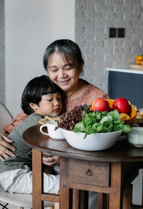 Positive senior ethnic female and cute little grandson hugging each other while having breakfast together at table with healthy salad and fruits in sunny morning